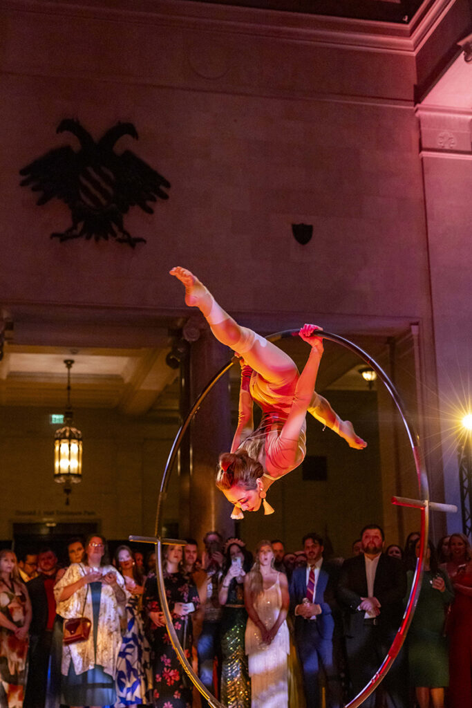 An acrobatic performer hangs inside of a large hoop, doing the splits upside down as a crowd watches at Party Arty: Painted Night.