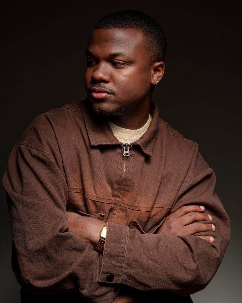 Malek Azrael crosses his arms and leans to the side in a dimly-lit monochromatic portrait.