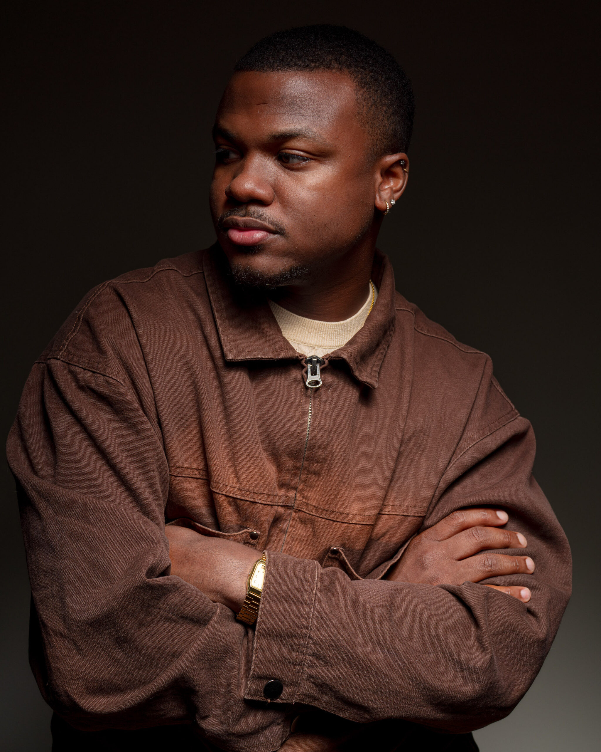 Malek Azrael crosses his arms and leans to the side in a dimly-lit monochromatic portrait.