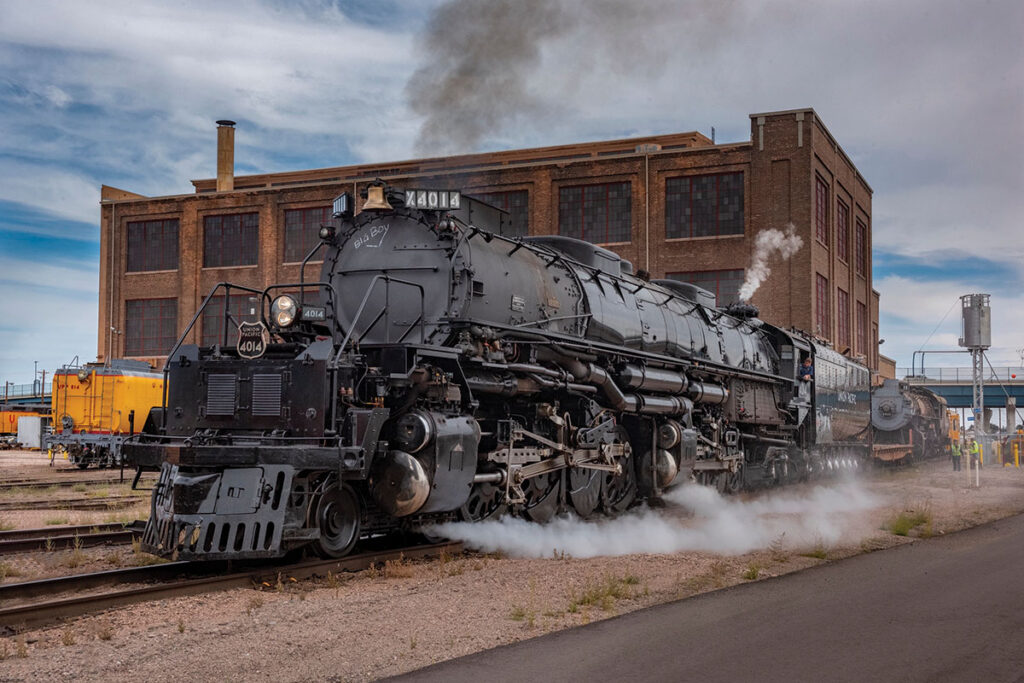 Big Boy number 1014, a large black steam locomotive sits on railroad tracks outside a historic brick rail building, with white steam billowing around its wheels.