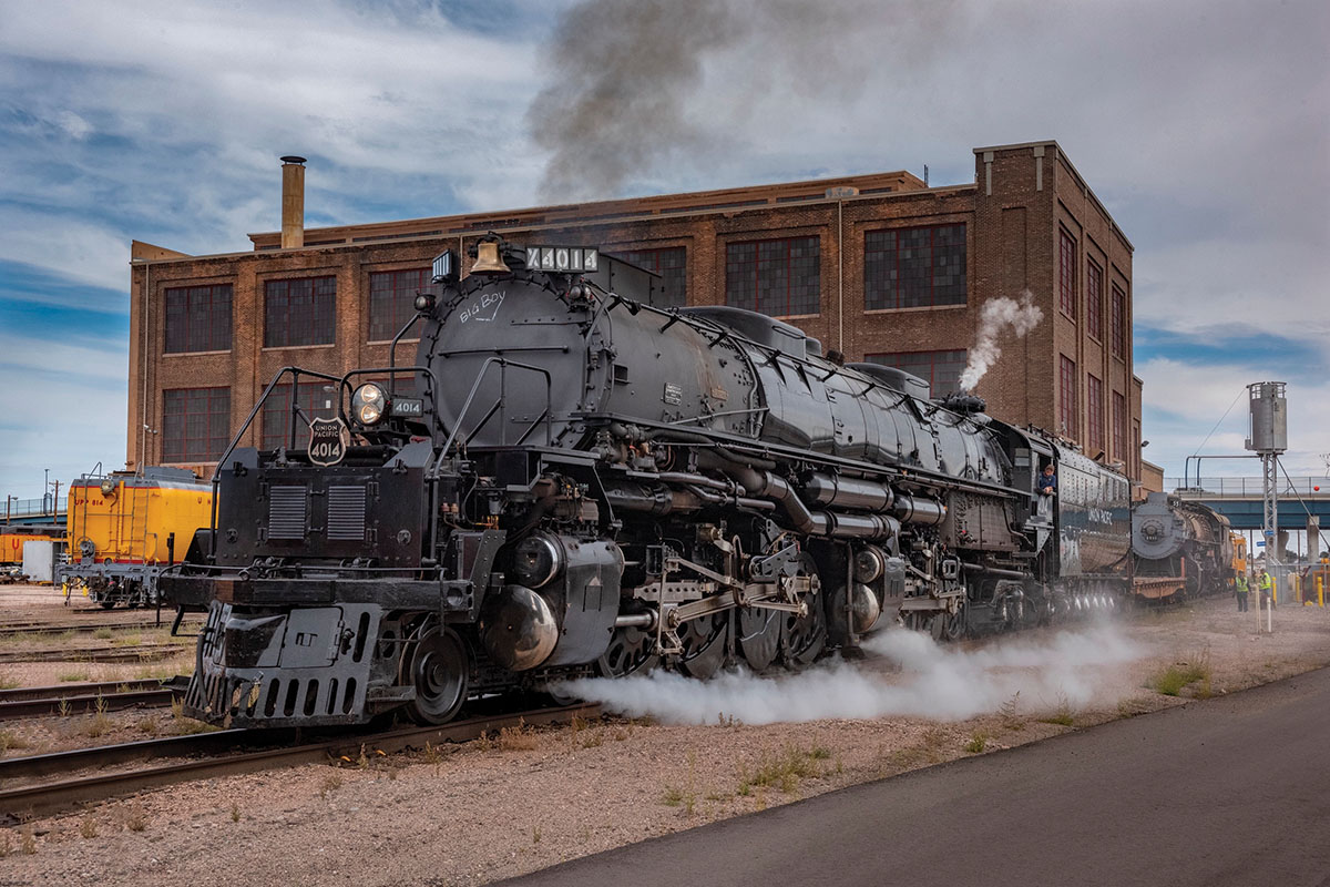Big Boy number 1014, a large black steam locomotive sits on railroad tracks outside a historic brick rail building, with white steam billowing around its wheels.